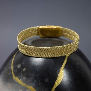 Op de afbeelding: Een gouden armband met een geweven ontwerp en een sluiting. De armband ligt op een zwart oppervlak met gouden accenten.