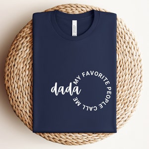 Könnte beinhalten: Ein marineblaues T-Shirt mit weißem Text, der "Meine Lieblingsmenschen nennen mich Dada" lautet.