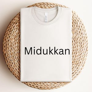 Könnte beinhalten: Weißes T-Shirt mit dem Text "Midukkan" in Schwarz auf der Vorderseite. Das T-Shirt liegt flach auf einer geflochtenen Strohoberfläche.
