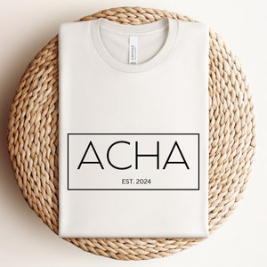 May include: White t-shirt with a black rectangle containing the text "ACHA EST. 2024" printed on the front.