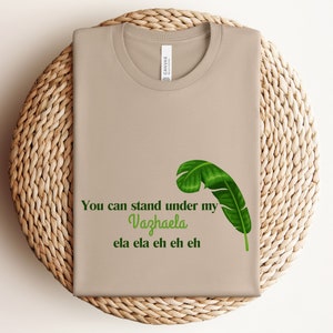 Puede incluir: Camiseta color beige con el texto verde "You can stand under my Vazhaela ela ela eh eh eh" y una ilustración de hoja de plátano verde. La camiseta está doblada sobre una superficie redonda y tejida.