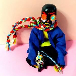 May include: A handcrafted wooden doll with a black head and a painted face featuring red and yellow details. The doll is dressed in a blue jacket, a yellow shirt, and a colourful braided scarf. A matching charm hangs from a blue cord.
