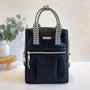 Könnte beinhalten: Schwarzer Rucksack mit Chevron-Muster an Griff und Trägern. Die Tasche hat einen goldenen Reißverschluss und eine kleine rechteckige Tasche auf der Vorderseite. Der Text "COURAGE" ist auf einem kleinen Banner auf der Vorderseite aufgedruckt.