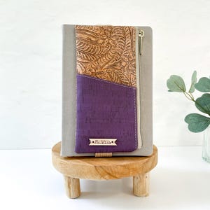 Peut inclure: Une pochette zippée en tissu de liège de couleur violette et marron avec une fermeture éclair dorée et une étiquette en métal sur laquelle est inscrit "carry courage".
