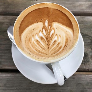 May include: A white ceramic mug filled with a latte, featuring a delicate, intricate foam design in the shape of a flower.