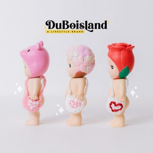 以下が含まれることがあります： 豚、花、バラの3つの異なる頭飾りをつけた小さなフィギュア。各フィギュアには小さなかぎ針編みのバッグが付いています。上部にはDuBoislandのロゴがあります。フィギュアは白い背景にあります。