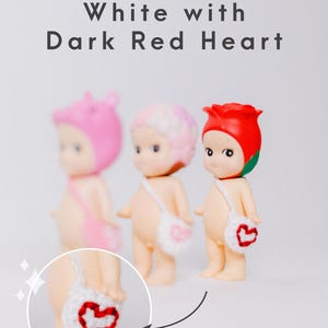 以下が含まれることがあります： ピンク、白、赤の帽子をかぶった3つの小さなフィギュア。各フィギュアは、赤いハートのデザインの小さな白いバッグを持っています。画像の上部には「White with Dark Red Heart」というテキストがあります。
