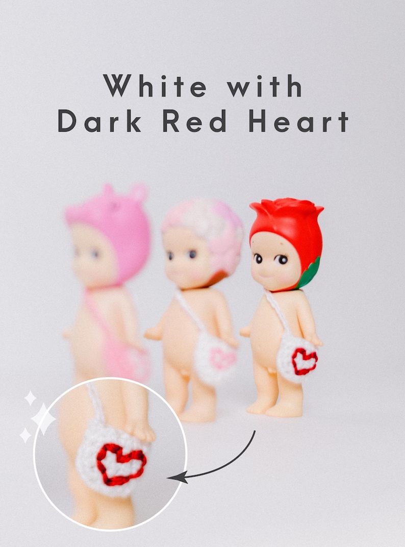 以下が含まれることがあります： ピンク、白、赤の帽子をかぶった3つの小さなフィギュア。各フィギュアは、赤いハートのデザインの小さな白いバッグを持っています。画像の上部には「White with Dark Red Heart」というテキストがあります。