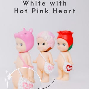 以下が含まれることがあります： ピンクのクマの帽子、ピンクと白の花柄、赤いバラなど、さまざまなヘッドカバーをかぶった3つの小さなフィギュア。 各フィギュアには、ピンクのハートのデザインが入った白いかぎ針編みのバッグが付いています。 上部には「White with Hot Pink Heart」という文字があります。