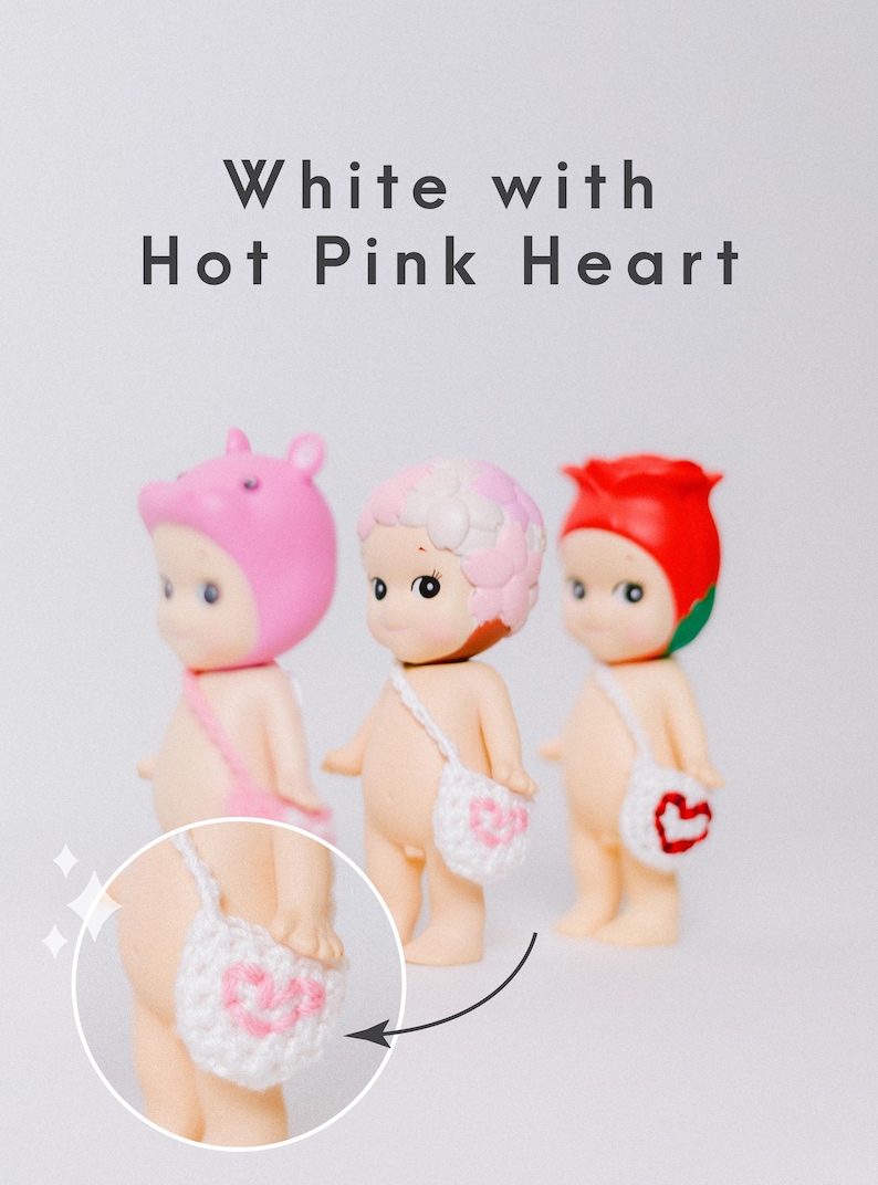 以下が含まれることがあります： ピンクのクマの帽子、ピンクと白の花柄、赤いバラなど、さまざまなヘッドカバーをかぶった3つの小さなフィギュア。 各フィギュアには、ピンクのハートのデザインが入った白いかぎ針編みのバッグが付いています。 上部には「White with Hot Pink Heart」という文字があります。