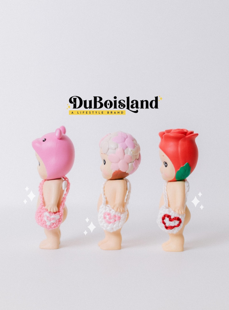 以下が含まれることがあります： 豚、花、バラの3つの異なる頭飾りをつけた小さなフィギュア。各フィギュアには小さなかぎ針編みのバッグが付いています。上部にはDuBoislandのロゴがあります。フィギュアは白い背景にあります。