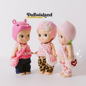 以下が含まれることがあります： ピンクのかぎ針編みのトップスとマッチするハンドバッグを着た3つの小さな人形。人形は、カバ、花、桃など、さまざまな帽子をかぶっています。人形はデニムのジーンズ、ヒョウ柄のパンツ、フローラルパンツを着用しています。背景にはDuBoislandのロゴがあります。