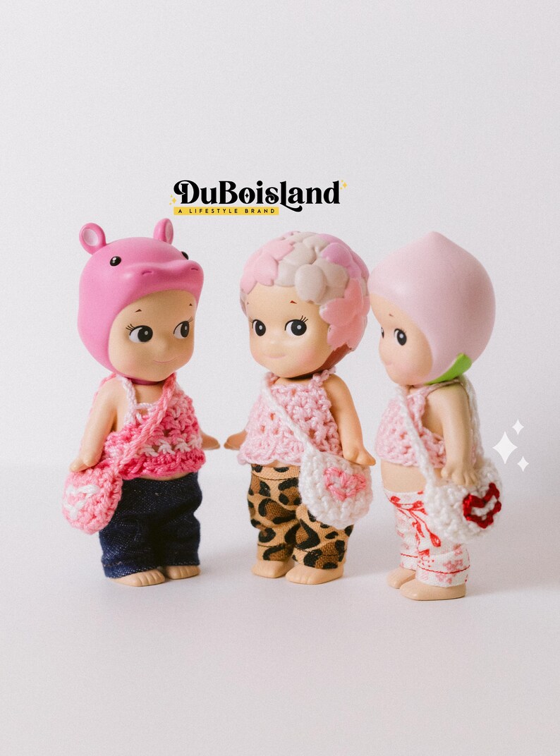 以下が含まれることがあります： ピンクのかぎ針編みのトップスとマッチするハンドバッグを着た3つの小さな人形。人形は、カバ、花、桃など、さまざまな帽子をかぶっています。人形はデニムのジーンズ、ヒョウ柄のパンツ、フローラルパンツを着用しています。背景にはDuBoislandのロゴがあります。