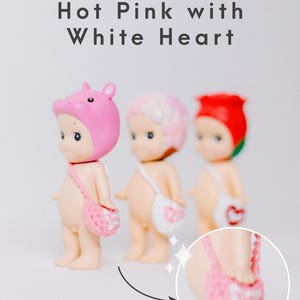 以下が含まれることがあります： さまざまな帽子と小さなかぎ針編みのバッグが付いた3つの小さな置物。上部には「Hot Pink with White Heart」という文字があります。バッグには白いハートのデザインがあります。1つの置物はピンクの豚の帽子をかぶり、別の置物はピンクと白の髪をしており、最後の置物は赤いバラを持っています。