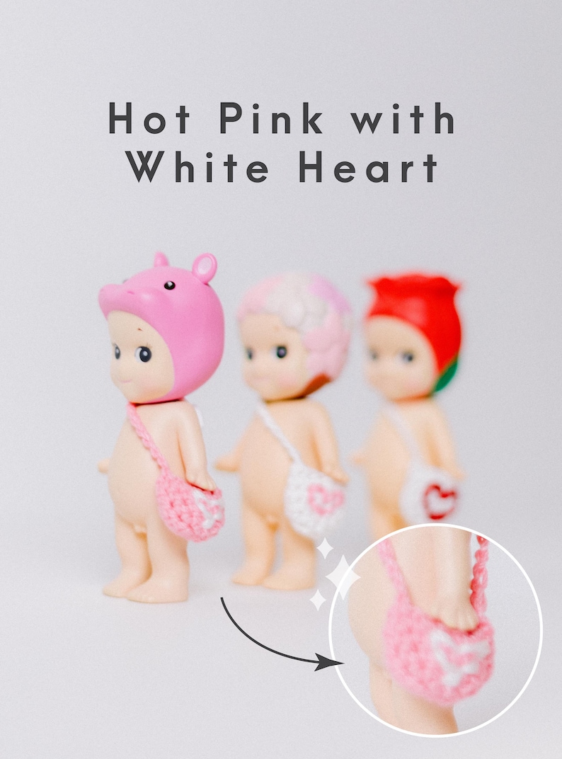 以下が含まれることがあります： さまざまな帽子と小さなかぎ針編みのバッグが付いた3つの小さな置物。上部には「Hot Pink with White Heart」という文字があります。バッグには白いハートのデザインがあります。1つの置物はピンクの豚の帽子をかぶり、別の置物はピンクと白の髪をしており、最後の置物は赤いバラを持っています。
