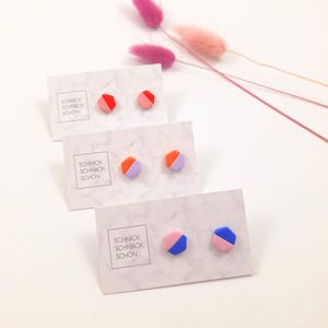 Könnte beinhalten: Drei Paar geometrische Ohrstecker in Rot, Rosa, Orange, Lila, Blau und Rosa. Jedes Paar wird auf einer weißen Karte mit dem Text "SCHNICK SCHNACK SCHON" präsentiert. Rosa und lila Trockenblumen sind im Hintergrund.