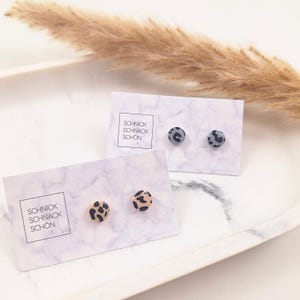 Peut inclure: Deux paires de boucles d'oreilles à clous avec des motifs animaliers sont présentées sur des cartes à motifs marbrés. Une paire présente des taches noires et beiges sur fond beige, l'autre des taches grises sur fond gris. Les cartes indiquent "SCHNICK SCHNACK SCHÖN".