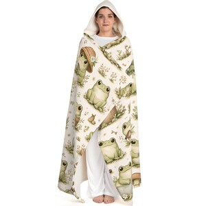 Peut inclure: Une couverture &agrave; capuche avec un motif r&eacute;p&eacute;t&eacute; de grenouilles de dessin anim&eacute; dans un cadre naturel. Les grenouilles sont de diff&eacute;rentes nuances de vert, certaines portant des chapeaux. Le fond est de couleur cr&egrave;me avec des &eacute;l&eacute;ments floraux et v&eacute;g&eacute;taux.