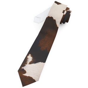 May include: A necktie with a cowhide pattern. The tie features a repeating pattern of brown, black, and white patches, resembling a cow's coat. The tie is angled, with the white fabric of the packaging visible.