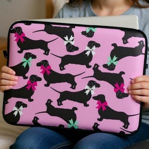 Op de afbeelding: Een roze laptoptas met een herhaald patroon van zwarte teckels met roze, witte en groene strikken. De hoes wordt vastgehouden door een persoon die met een laptop op een bed zit.