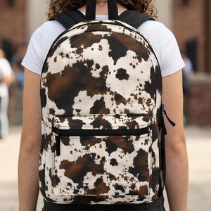 May include: A backpack with a cowhide pattern in shades of brown, black, and white. The backpack has a front pocket, black straps, and a black top handle. The person is wearing a white t-shirt.