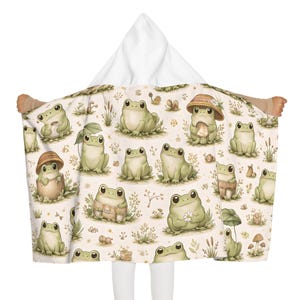 Peut inclure: Une serviette &agrave; capuche avec un motif fantaisiste de grenouilles vert clair dans diverses poses, certaines portant des chapeaux ou tenant des objets. Le fond est de couleur cr&egrave;me avec des illustrations florales et v&eacute;g&eacute;tales. La capuche est blanche.
