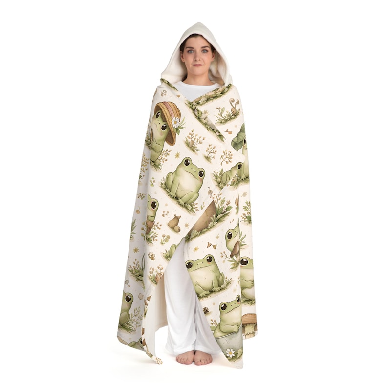 Peut inclure: Une couverture &agrave; capuche avec un motif r&eacute;p&eacute;t&eacute; de grenouilles de dessin anim&eacute; dans un cadre naturel. Les grenouilles sont de diff&eacute;rentes nuances de vert, certaines portant des chapeaux. Le fond est de couleur cr&egrave;me avec des &eacute;l&eacute;ments floraux et v&eacute;g&eacute;taux.