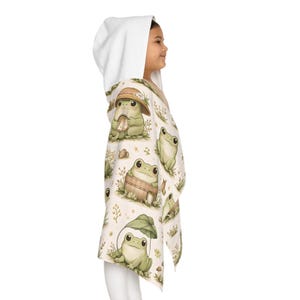 Peut inclure: Une serviette &agrave; capuche cr&egrave;me orn&eacute;e d'un motif de grenouilles. Les grenouilles sont repr&eacute;sent&eacute;es dans diverses poses, certaines portant des chapeaux ou tenant des champignons. La capuche est blanche.
