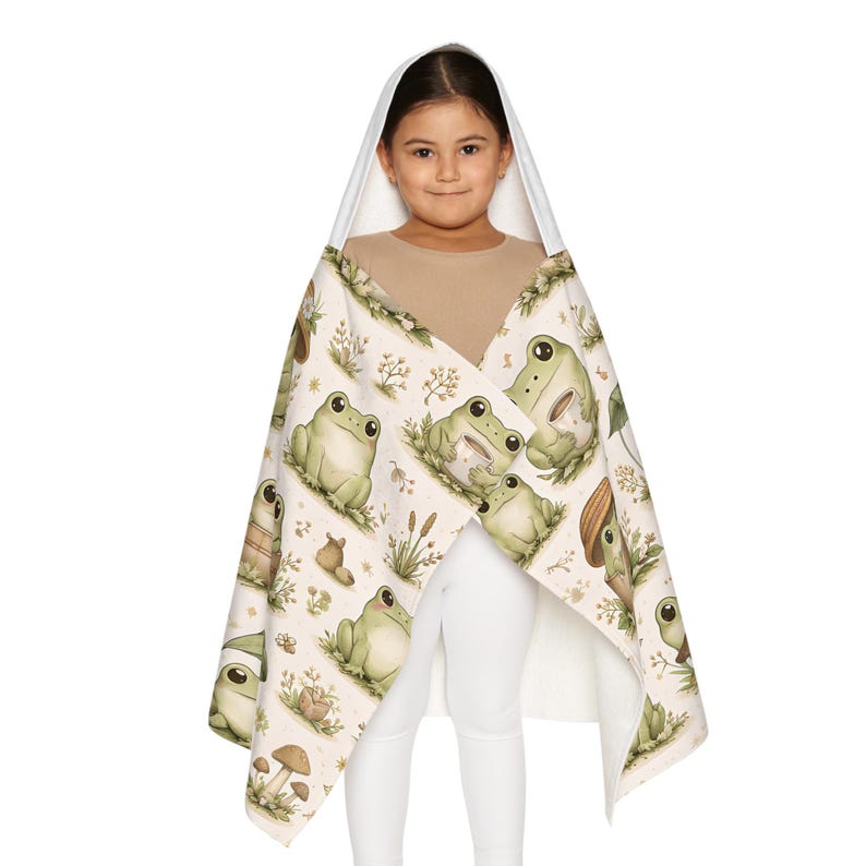Peut inclure: Une serviette &agrave; capuche avec un motif de grenouilles vertes, beiges et marron. La serviette a une capuche blanche et est enroul&eacute;e autour d'une personne. Le motif comprend des illustrations de grenouilles, de champignons et de plantes.