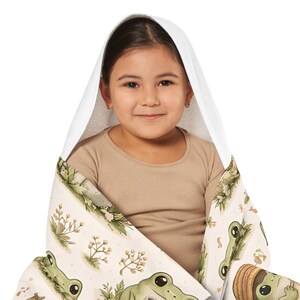 Peut inclure: Un enfant envelopp&eacute; dans une serviette &agrave; capuche blanche avec un motif fantaisiste de grenouilles et de plantes. La serviette pr&eacute;sente un fond beige clair avec des illustrations de grenouilles vertes et des accents botaniques.