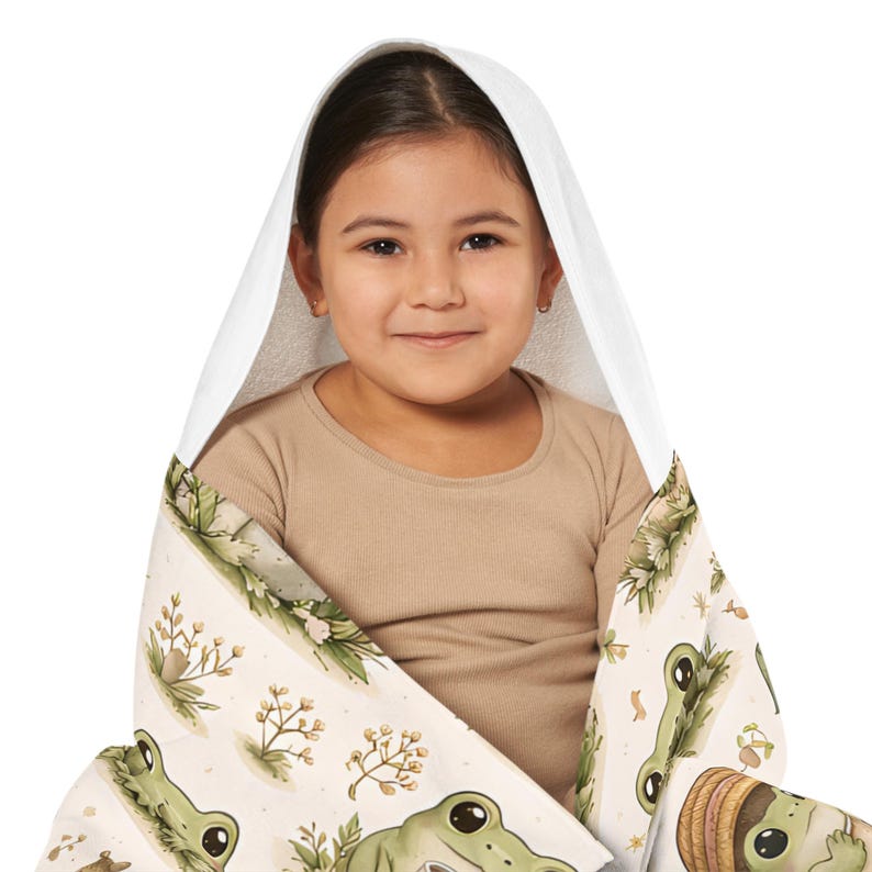Peut inclure: Un enfant envelopp&eacute; dans une serviette &agrave; capuche blanche avec un motif fantaisiste de grenouilles et de plantes. La serviette pr&eacute;sente un fond beige clair avec des illustrations de grenouilles vertes et des accents botaniques.