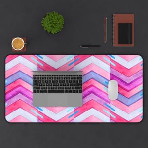 May include: A desk mat with a watercolor chevron pattern in shades of pink, purple, and blue. A laptop, mouse, pen, notebook, and a cup of coffee are on the mat. A small potted plant is also visible.