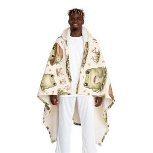 Peut inclure: Une couverture &agrave; capuche cr&egrave;me avec un motif de grenouilles vertes, de fleurs et de champignons. La couverture a une capuche doubl&eacute;e de sherpa blanc. La personne porte un pantalon blanc et un t-shirt blanc.