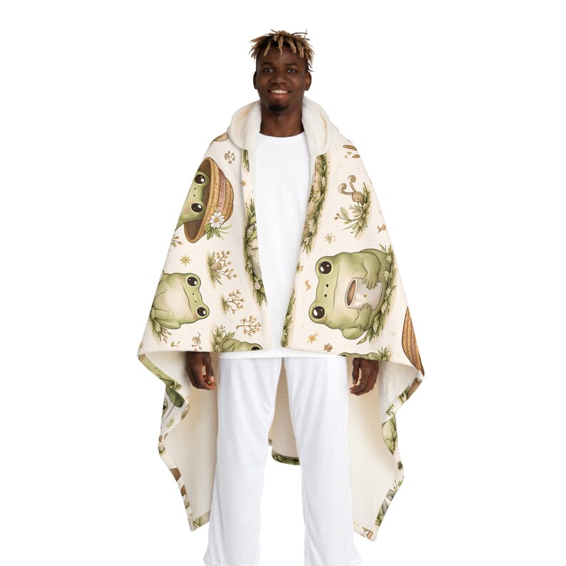 Peut inclure: Une couverture &agrave; capuche cr&egrave;me avec un motif de grenouilles vertes, de fleurs et de champignons. La couverture a une capuche doubl&eacute;e de sherpa blanc. La personne porte un pantalon blanc et un t-shirt blanc.