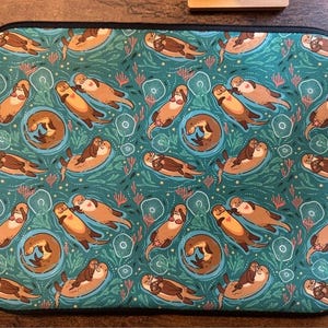 May include: A teal laptop sleeve featuring a repeating pattern of cartoon otters swimming in a sea of teal and green. The otters are brown and tan, with some cuddling. The design includes coral and other aquatic elements.