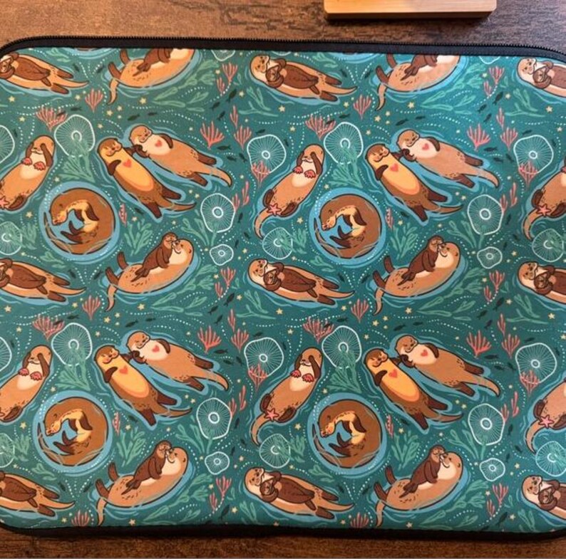 May include: A teal laptop sleeve featuring a repeating pattern of cartoon otters swimming in a sea of teal and green. The otters are brown and tan, with some cuddling. The design includes coral and other aquatic elements.