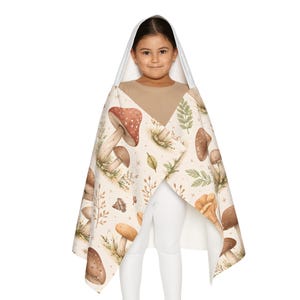 Peut inclure: Une serviette &agrave; capuche avec un motif de champignons marron, beige et vert. La serviette a une capuche blanche et un design enveloppant. Le motif pr&eacute;sente des illustrations de diff&eacute;rents champignons et feuilles. Parfait pour s&eacute;cher apr&egrave;s le bain ou la baignade.