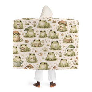Peut inclure: Une couverture &agrave; capuche avec un motif r&eacute;p&eacute;t&eacute; de grenouilles de dessin anim&eacute; dans diverses poses, certaines portant des chapeaux ou tenant des objets. Les grenouilles sont dans des tons de vert et de beige, sur un fond cr&egrave;me avec des accents floraux et de champignons.