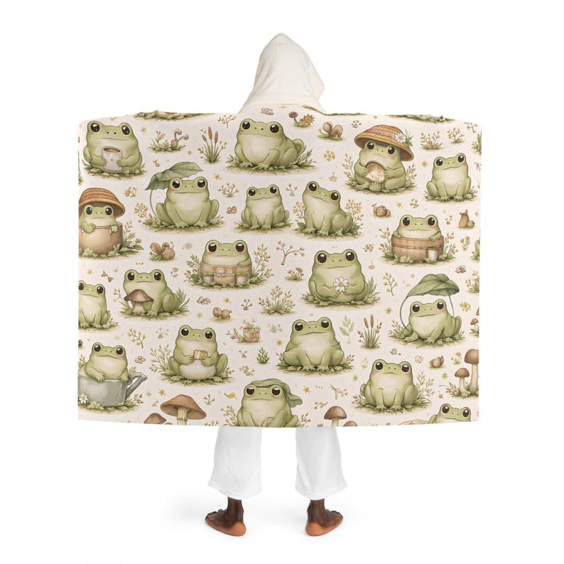 Peut inclure: Une couverture &agrave; capuche avec un motif r&eacute;p&eacute;t&eacute; de grenouilles de dessin anim&eacute; dans diverses poses, certaines portant des chapeaux ou tenant des objets. Les grenouilles sont dans des tons de vert et de beige, sur un fond cr&egrave;me avec des accents floraux et de champignons.
