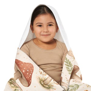 May include: A hooded towel with a mushroom design. The towel has a white hood and a beige background with illustrations of mushrooms, leaves, and other botanical elements. The towel is wrapped around a person.