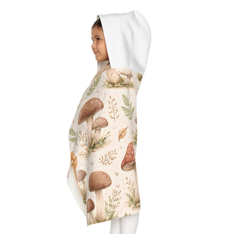 May include: A hooded towel with a pattern of mushrooms and foliage in brown, beige, and green. The towel has a white hood and is wrapped around a person. The design includes various mushroom illustrations and botanical elements.