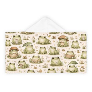 Peut inclure: Une serviette &agrave; capuche avec un motif r&eacute;p&eacute;t&eacute; de grenouilles de dessin anim&eacute; dans diverses poses, certaines portant des chapeaux ou tenant des objets. Les grenouilles sont vert clair avec des ventres blancs, sur un fond beige avec des accents floraux et de champignons.
