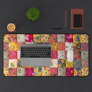 May include: A colorful patchwork desk mat with a laptop, mouse, and other office supplies. The mat features a variety of floral patterns in shades of pink, yellow, and green. A small potted plant, a cup of coffee, and a notebook are also visible.