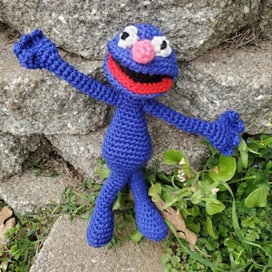 Könnte beinhalten: Handgemachte blaue Häkelpuppe mit rundem Körper, ausgestreckten Armen und ausgestreckten Beinen. Die Puppe hat eine rosa Nase, einen roten Mund und weiße und schwarze Augen. Die Puppe befindet sich auf einer Steinoberfläche mit Grün im Hintergrund.