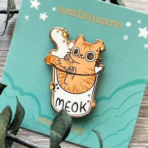 May include: Gold enamel pin featuring a cartoon orange cat with big eyes inside a white cup with the word "MEOK" written on it. The pin is on a teal blue card with white stars and the text "acatsquare enamel pin".