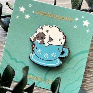 May include: A blue enamel pin featuring a cartoon sheep wearing a pink bow, sleeping in a blue teacup with white flowers. The pin is on a green card with the text "acatsquare" and "enamel pin".
