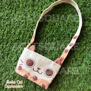 May include: Beige canvas Boba Cat cupholder with a cat face design. The cupholder has a brown and tan color scheme, with a matching strap featuring brown circles. The text "Boba Cat Cupholder" is printed on the bottom.