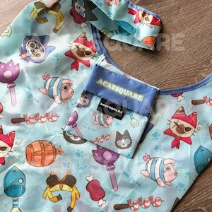 May include: Light blue tote bag with a repeating pattern of cartoon cats, food, and weapons. The bag has a small pocket with the text "ACATSQUARE". The bag is made of a lightweight material and is folded.