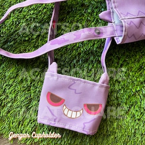 May include: A purple Gengar cup holder with a matching strap, featuring a smiling Gengar face with pink and brown accents. The cup holder is made of fabric and has a stitched design. The text "Gengar Cupholder" is at the bottom.