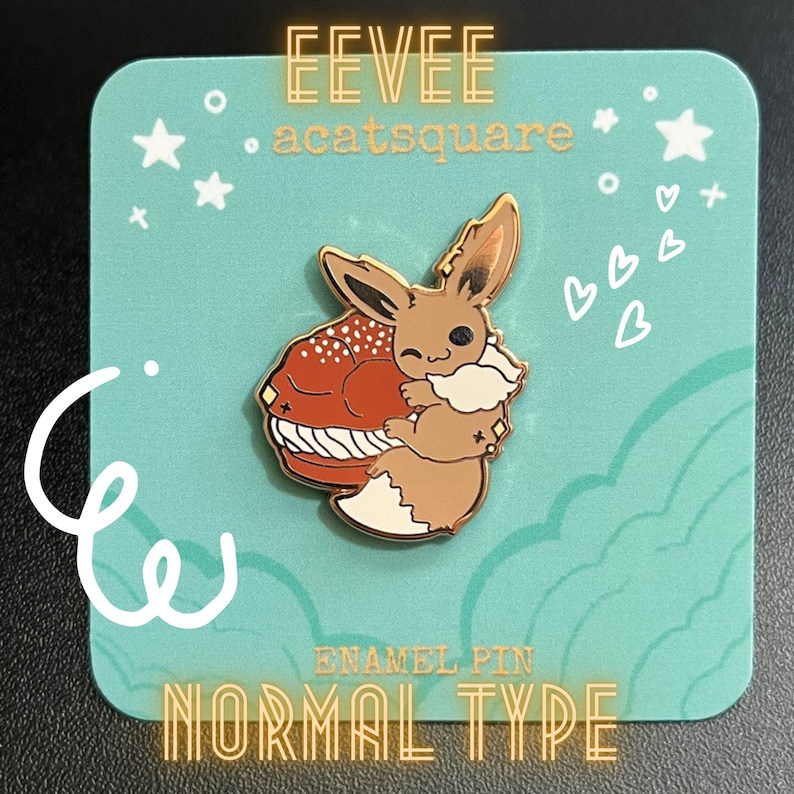 May include: An enamel pin featuring Eevee hugging a macaron. The pin is brown, white, and gold with the text "EEVEE" and "NORMAL TYPE". The pin is on a teal background with white stars and hearts.